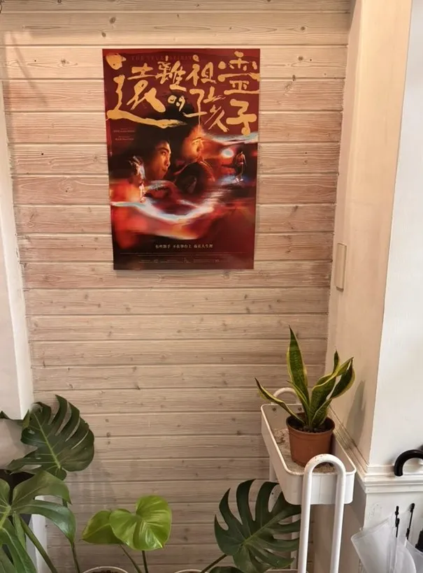 《遠離祖靈的孩子》的電影海報，掛在木板牆上，牆下擺著幾盆龜背芋與一盆虎尾蘭